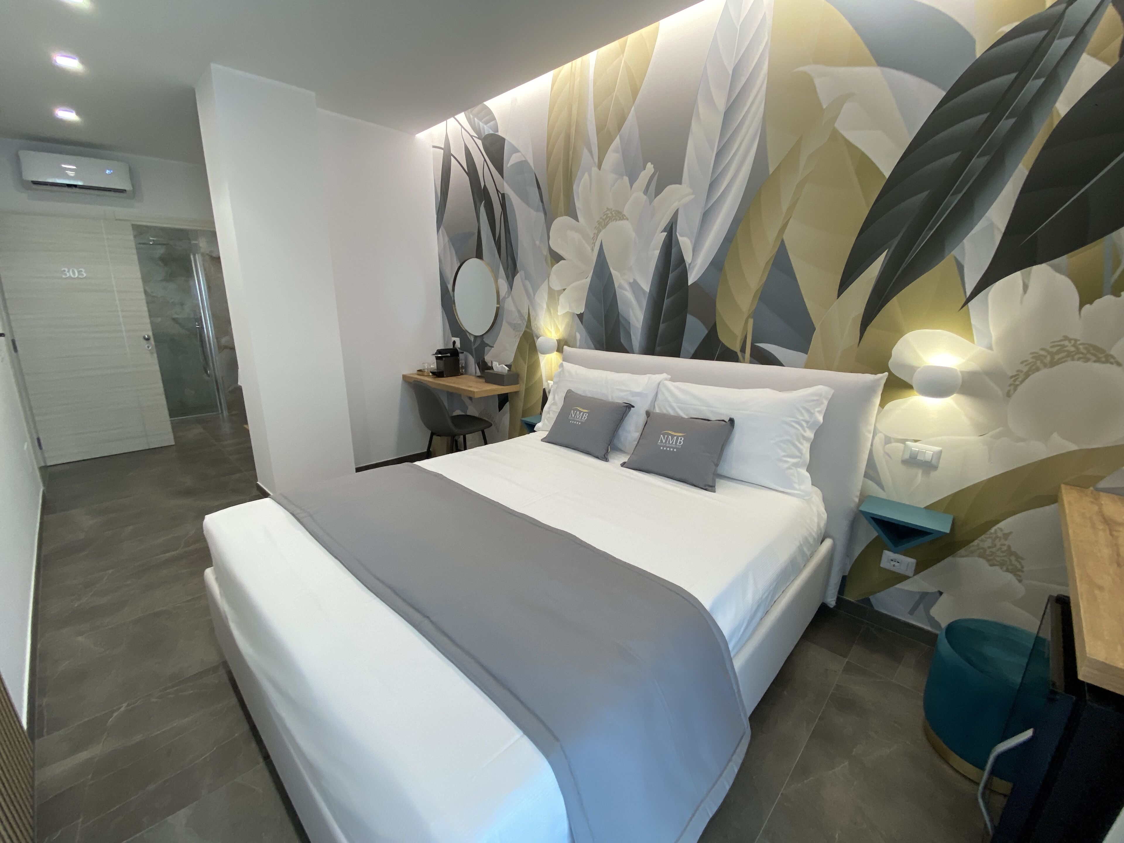 A modern bedroom with a large bed, a desk, and a chair. There is a mirror on the wall and a decorative wall panel.