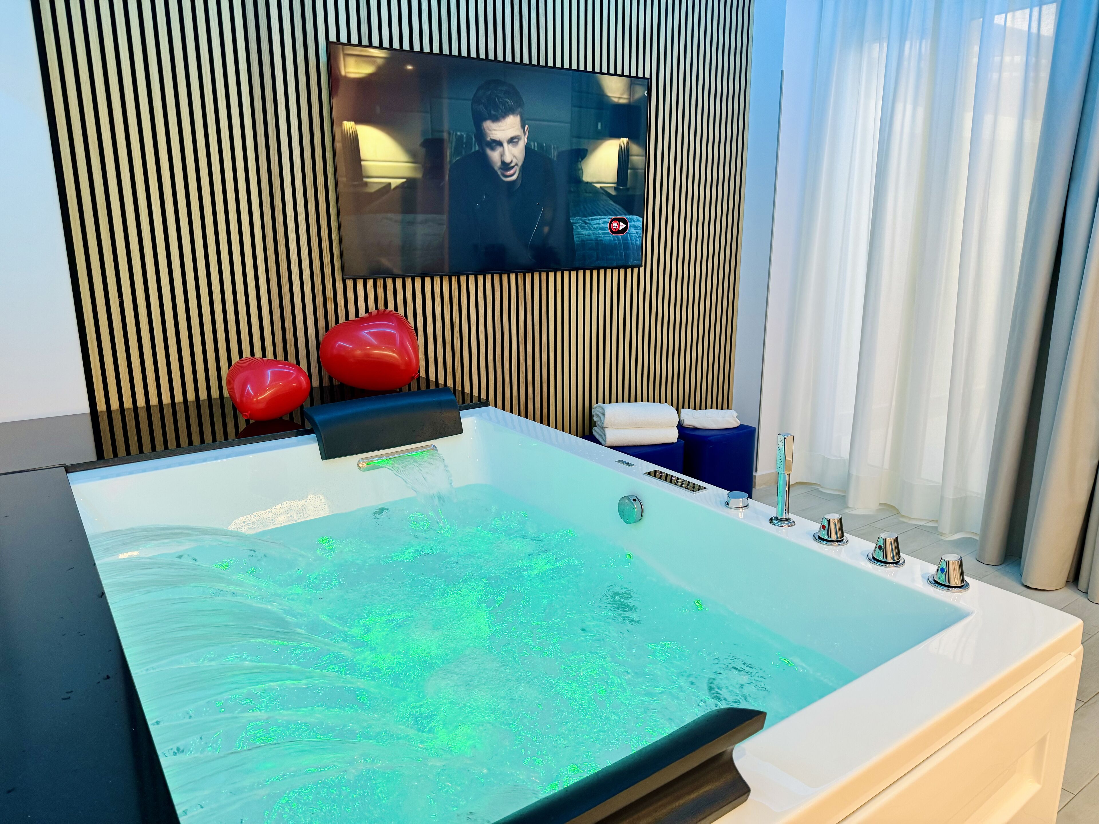 Un baño moderno con una bañera grande, un televisor montado y taburetes rojos con forma de corazón.