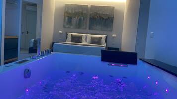 Une chambre moderne avec un grand lit, une table de chevet, un bureau et une grande baignoire dotée d’un éclairage violet.