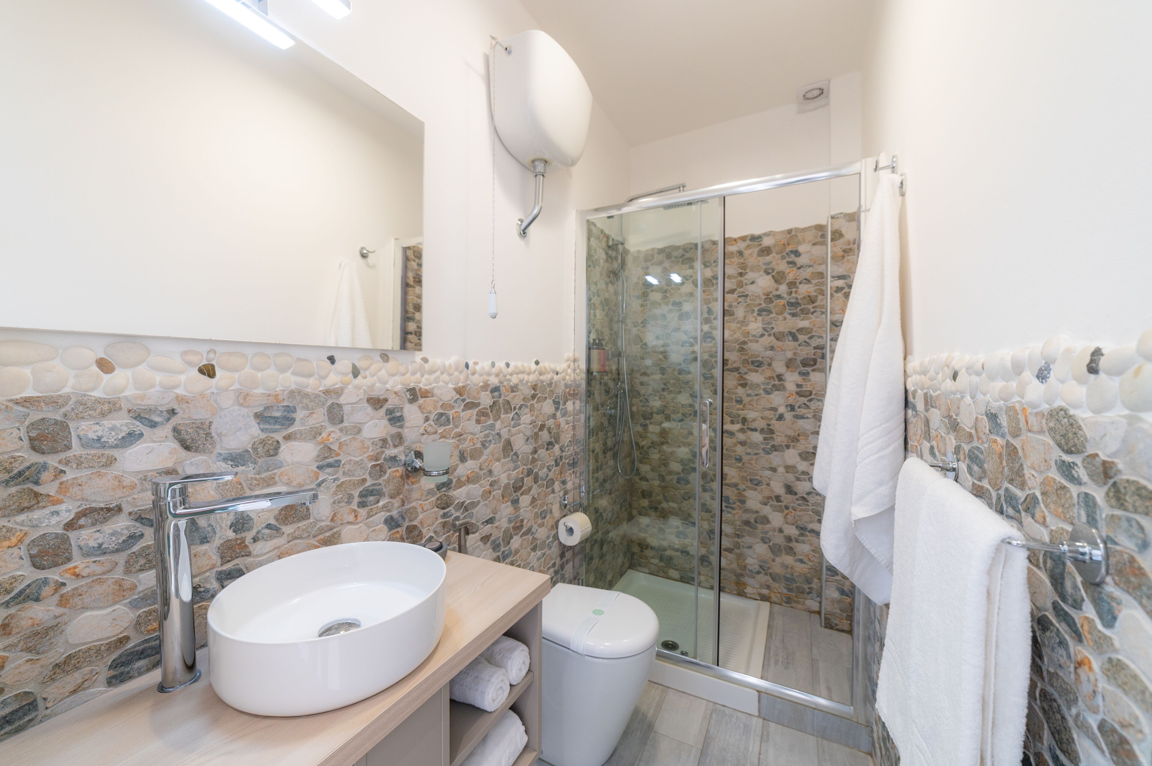 Une salle de bain avec un mur recouvert d’une mosaïque de pierre, un lavabo blanc, une douche en verre et un porte-serviettes.