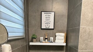 Un bagno con un WC, un asciugatore di mani, un cestino e uno scaffale con articoli da toilette.