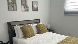 Un letto rifatto con una coperta a righe bianche e verdi, una testiera scura e due cuscini. Sopra il letto, tre elementi decorativi a parete, un comodino con una lampada e una finestra con persiane.