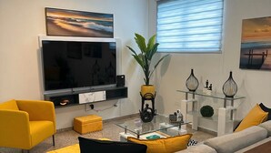 Un soggiorno moderno con un divano grigio, poltrone gialle, una TV a schermo piatto, un tavolino da caffè in vetro e una pianta in vaso.