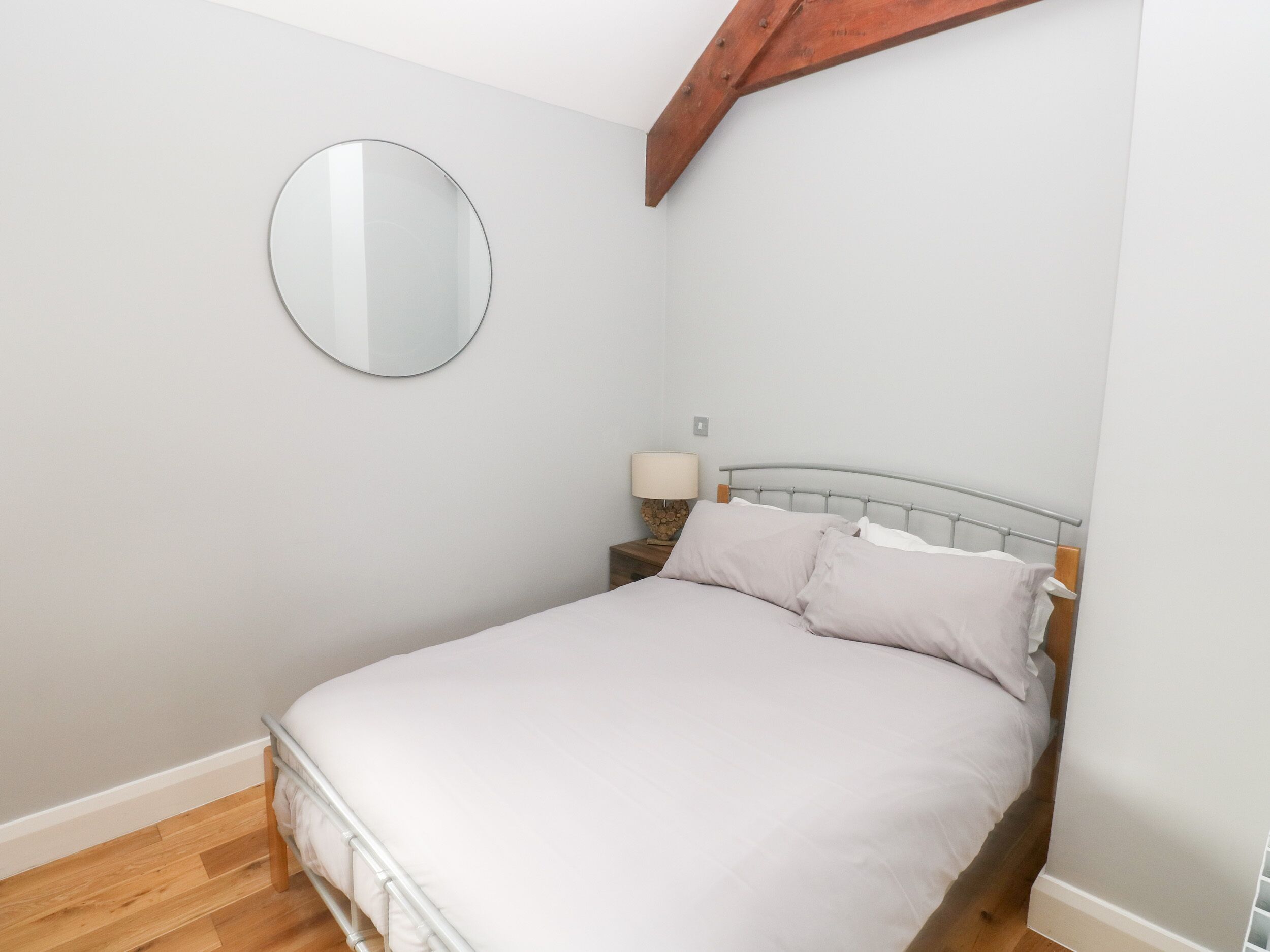 Un dormitorio con cama, un espejo redondo en la pared, una mesita de noche con lámpara y una viga de techo de madera.