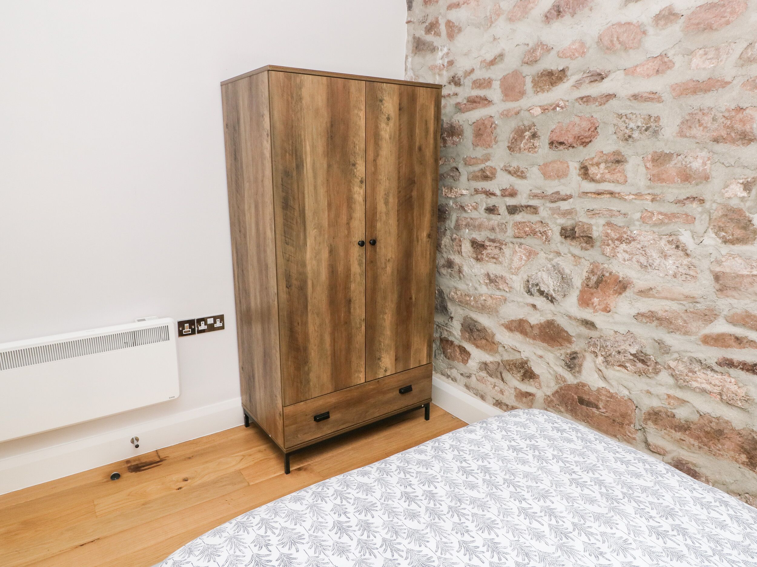 Een kamer met een houten garderobe, een radiator, een bakstenen muur en een bed met een decoratieve spreidek.