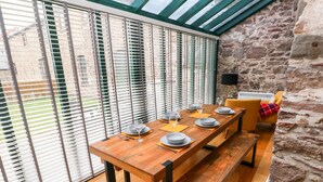 A dining area with a wooden table set for four, large windows with blinds, a stone wall, and a yellow armchair.