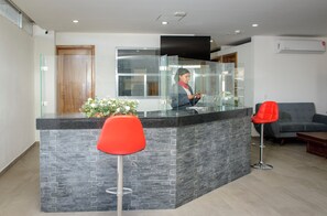 A reception area with a glass partition, a reception desk, a red chair, a grey sofa, and a wooden door. - MC HOTEL SALDARRIAGA QUERETARO (El Marques)