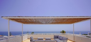 Ein überdachter Außenbereich mit Meerblick, ausgestattet mit einem Holzboden, weißen Möbeln und einer Pergola.