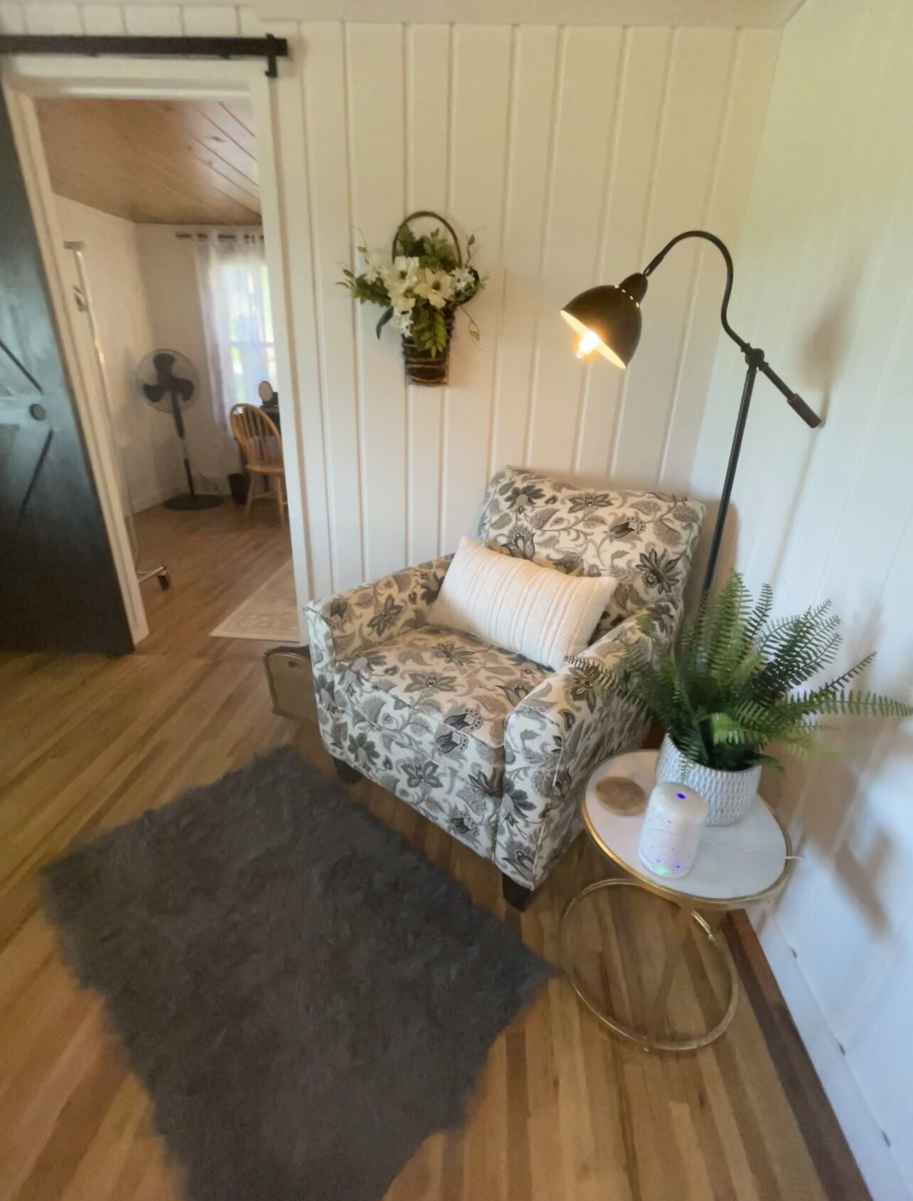 Een gezellige kamer met een gestreept fauteuil, een klein bijzettafeltje met een plant, een vloerlamp en een donker vloerkleed.
