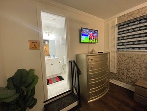 A hotel room with a television mounted on the wall, a dresser, a plant, and a door leading to a bathroom.