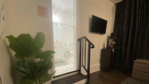 A living room with a large potted plant, a staircase leading to a bathroom, a mounted TV, and a dark curtain. - Magnolia Suite Vacation Home -Private Entry, Old Canton N. Jackson (Jackson)