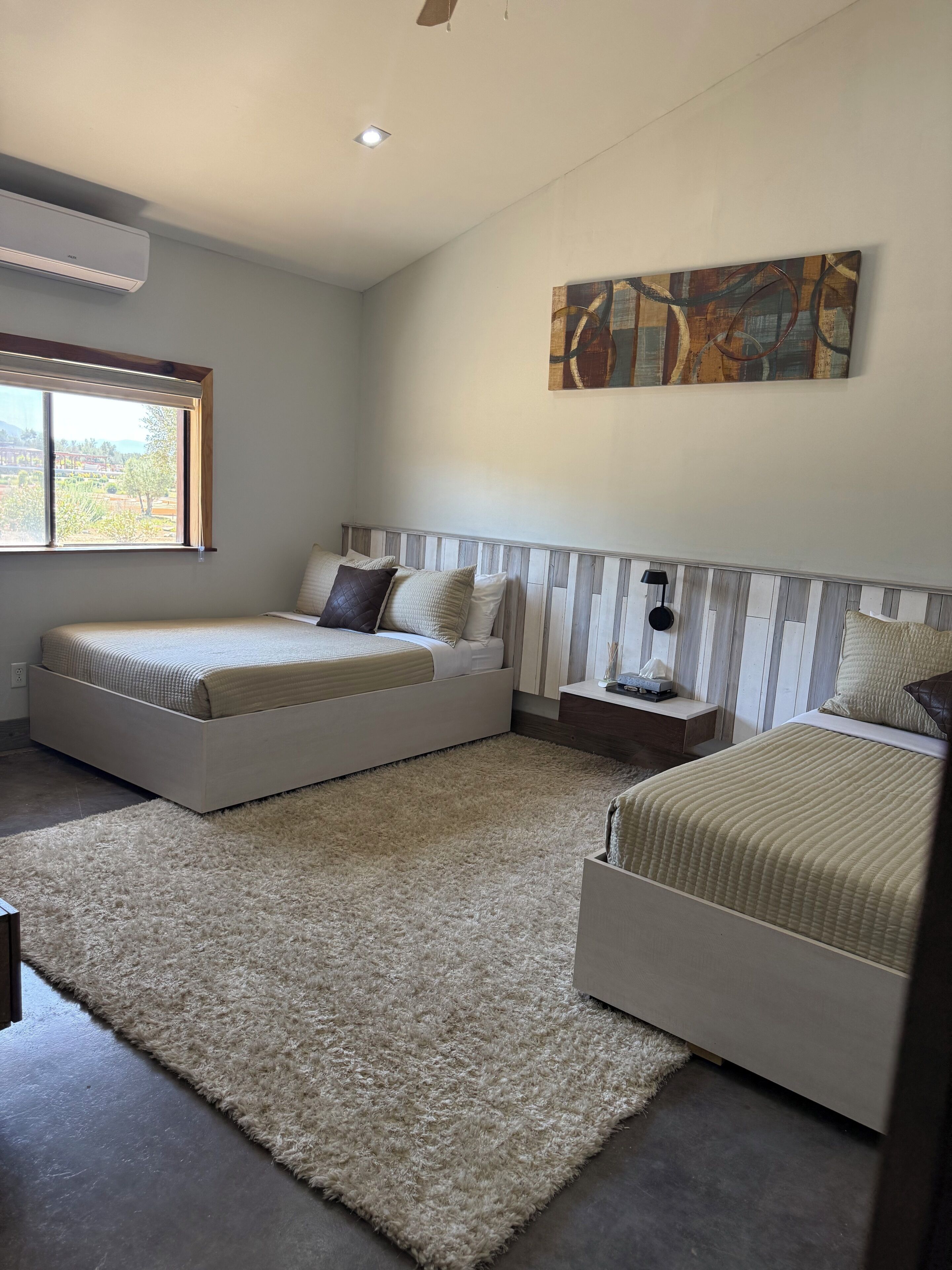 A bedroom with two beds, a large window, a painting, and a ceiling fan.