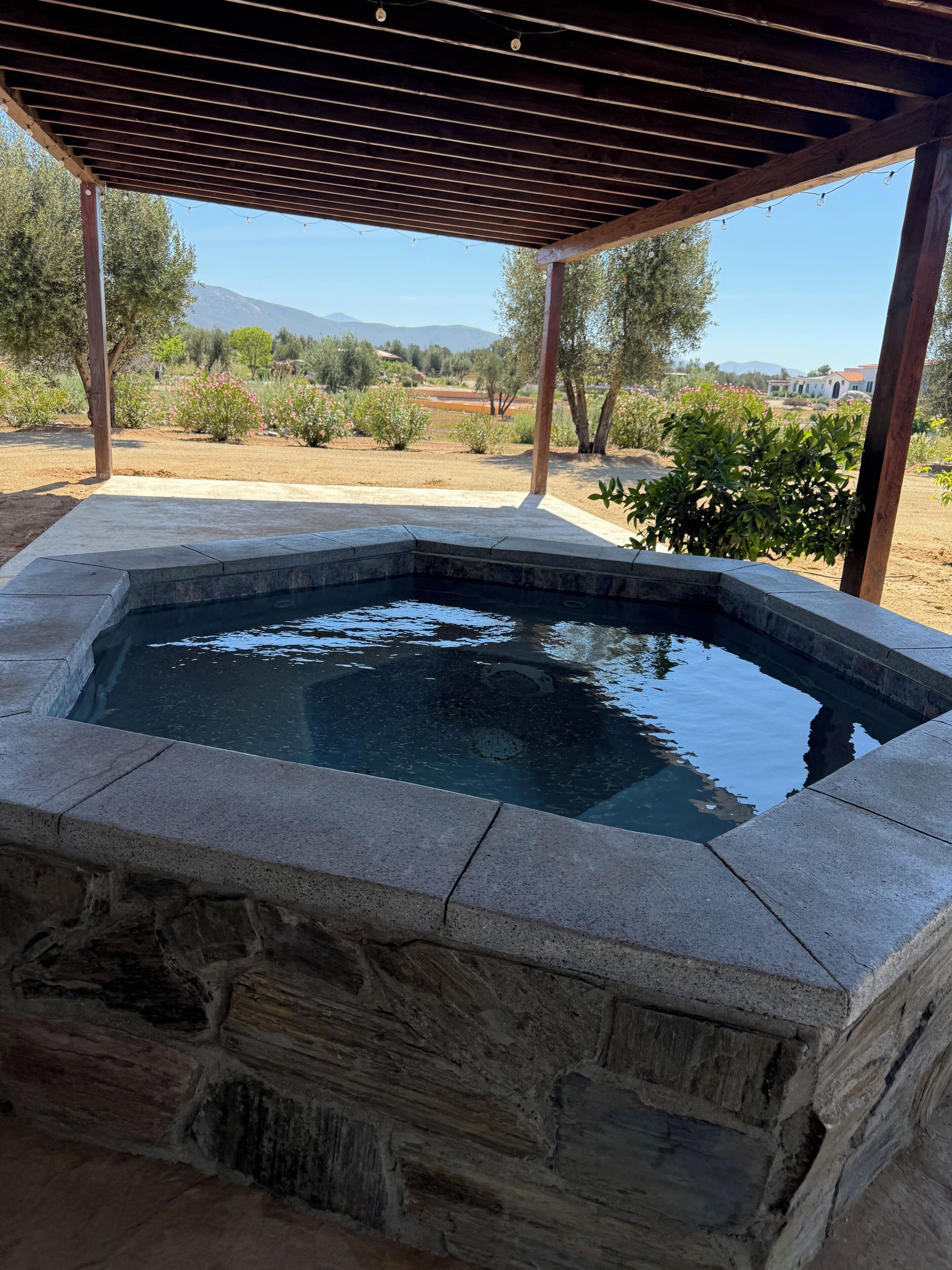 A stone hot tub under a wooden pergola with a view of a landscaped garden and distant mountains.