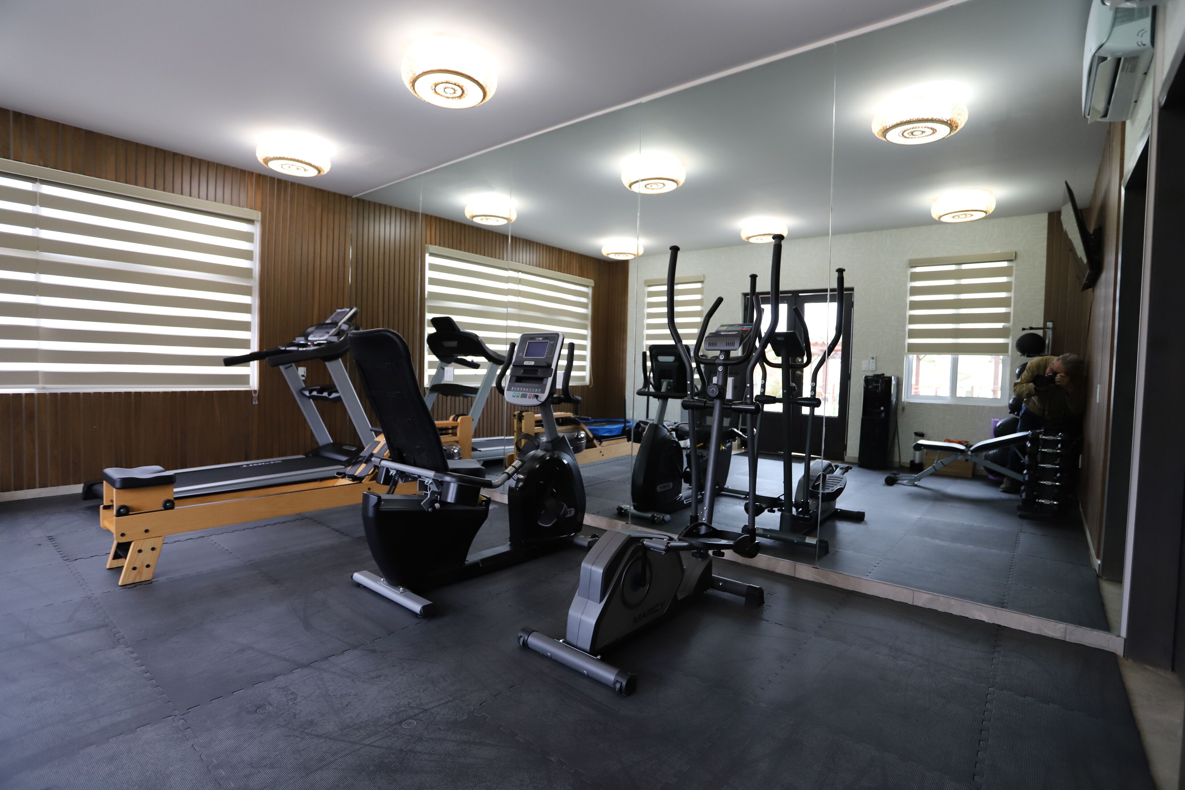 A gym with exercise equipment, large windows with blinds, and a mirror reflecting the equipment.