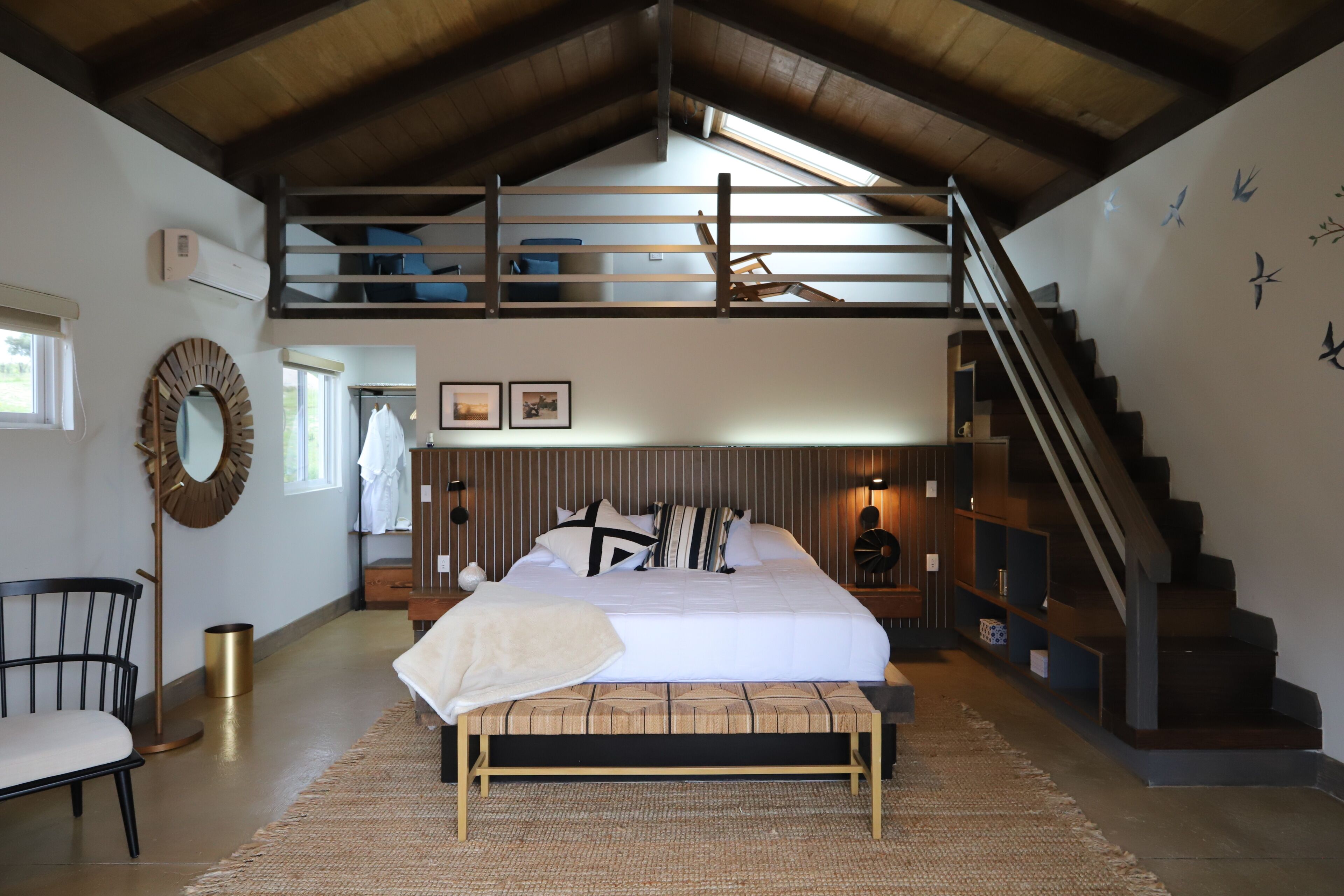 A bedroom with a loft bed, a bench, a staircase, and a wall-mounted mirror.