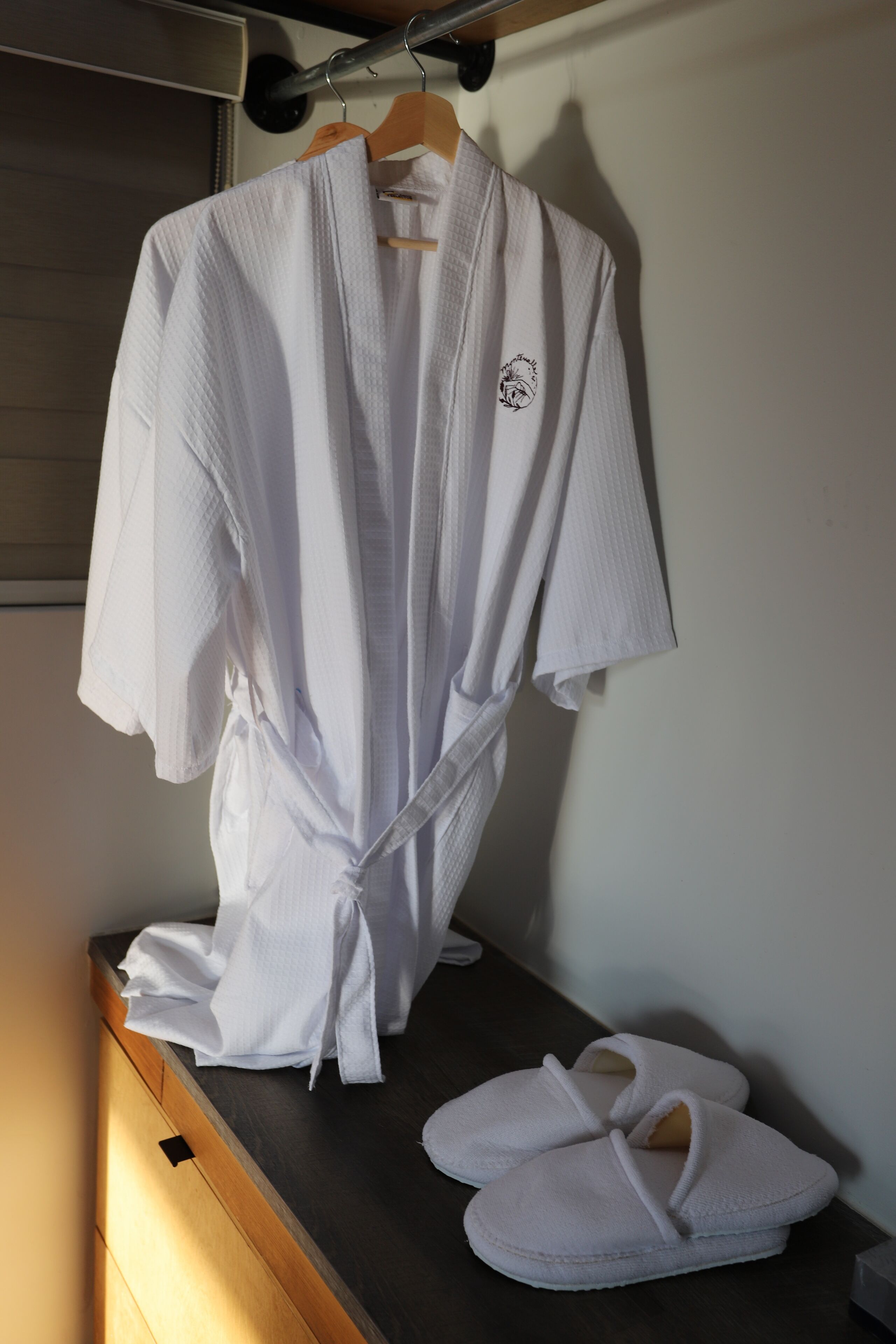 A white robe hanging on a wooden hanger and a pair of white slippers on a wooden surface.