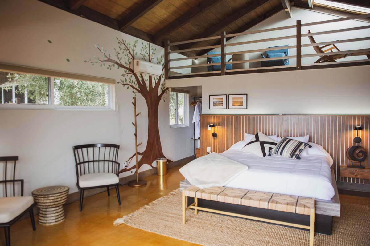 A bedroom with a bunk bed, a tree mural, a bedside table, a chair, and a window.