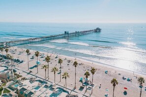 A beach with a long pier extending into the ocean, a pool area with lounge chairs, and palm trees lining the beach. - Hottest Neighborhood! Walk to Beach! (Oceanside)