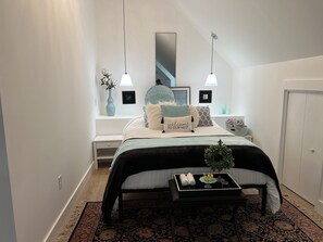 A bedroom with a bed, bedside table, nightstand, and a window with a view of a staircase.