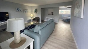 A modern living room with a grey sofa, a white coffee table, and a TV. There is a dining area in the background with a table and chairs. - NEW LISTING - 3 Min Walk-Beach, Hot Tub, Tennis, Wi-Fi (Riviera Beach)