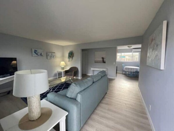 A modern living room with a grey sofa, a white coffee table, and a TV. There is a dining area in the background with a table and chairs.