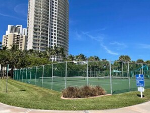 A tennis court with a green fence, surrounded by grass and palm trees, with a tall building in the background. - Hot Tub, Walk to Beach, Snorkeling, Surfing (Riviera Beach)