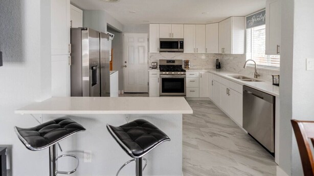 Una cocina moderna con armarios blancos, una isla central con dos taburetes y electrodomésticos de acero inoxidable, además de un ventanal con persianas.