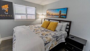 Un dormitorio con cama, una mesita de noche, una lámpara, una ventana con persianas, un cuadro de un paisaje urbano y un logotipo de Gucci en la pared.