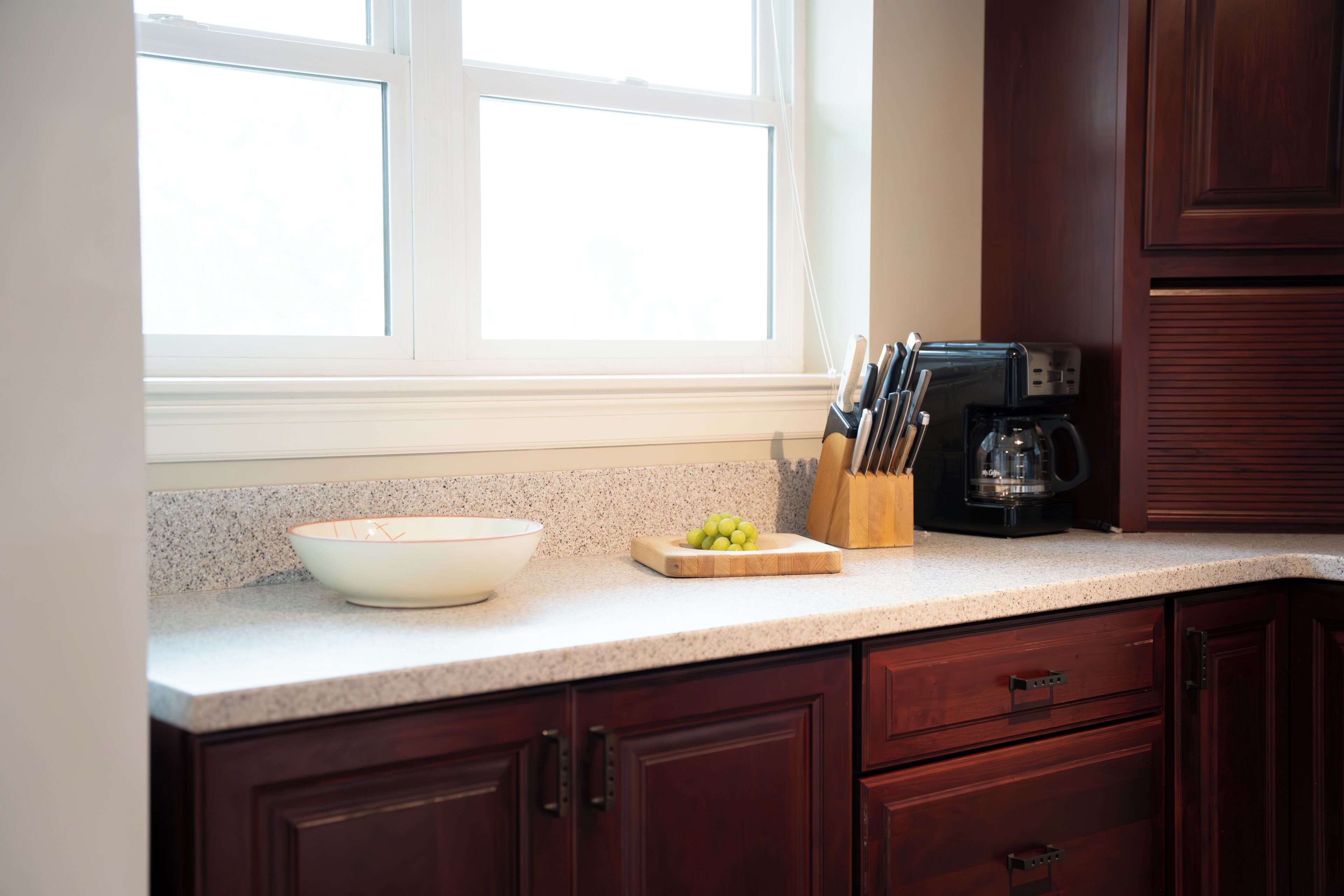 A kitchen with a granite countertop, a white bowl, a cutting board with grapes, a coffee maker, and wooden cabinets.