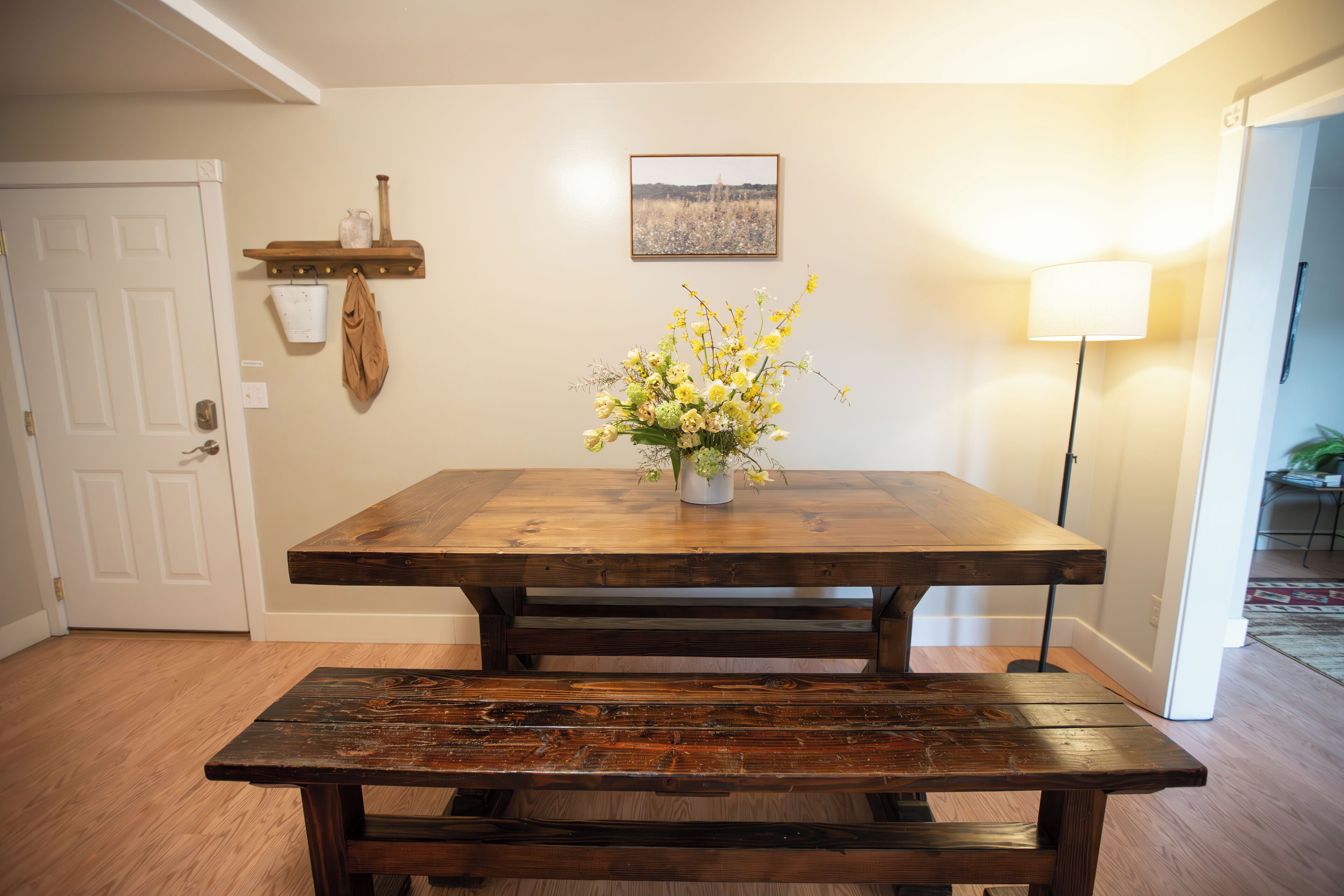 A wooden dining table with a bench, a vase of flowers, a wall-mounted shelf with a pitcher, and a framed picture.