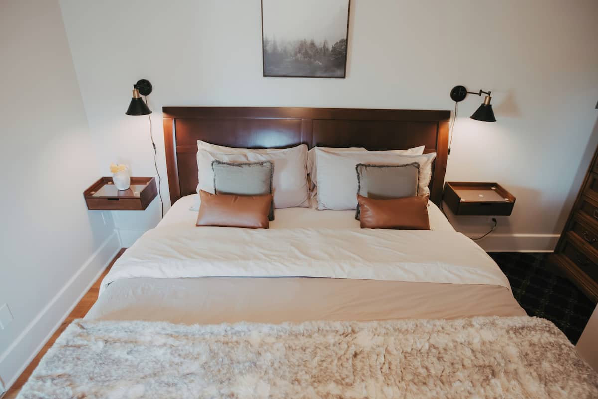 Una cama bien hecha con un edredón blanco esponjoso, dos mesitas de noche con lámparas y un cuadro enmarcado en la pared.
