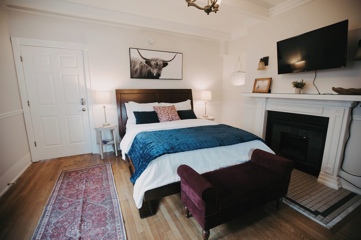 Un dormitorio con una cama grande, un televisor sobre la chimenea y una puerta a la izquierda.