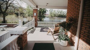 Eine überdachte Terrasse mit Blick auf einen Fluss, eine Ziegelwand, ein Holzstuhl und ein Blumentopf.