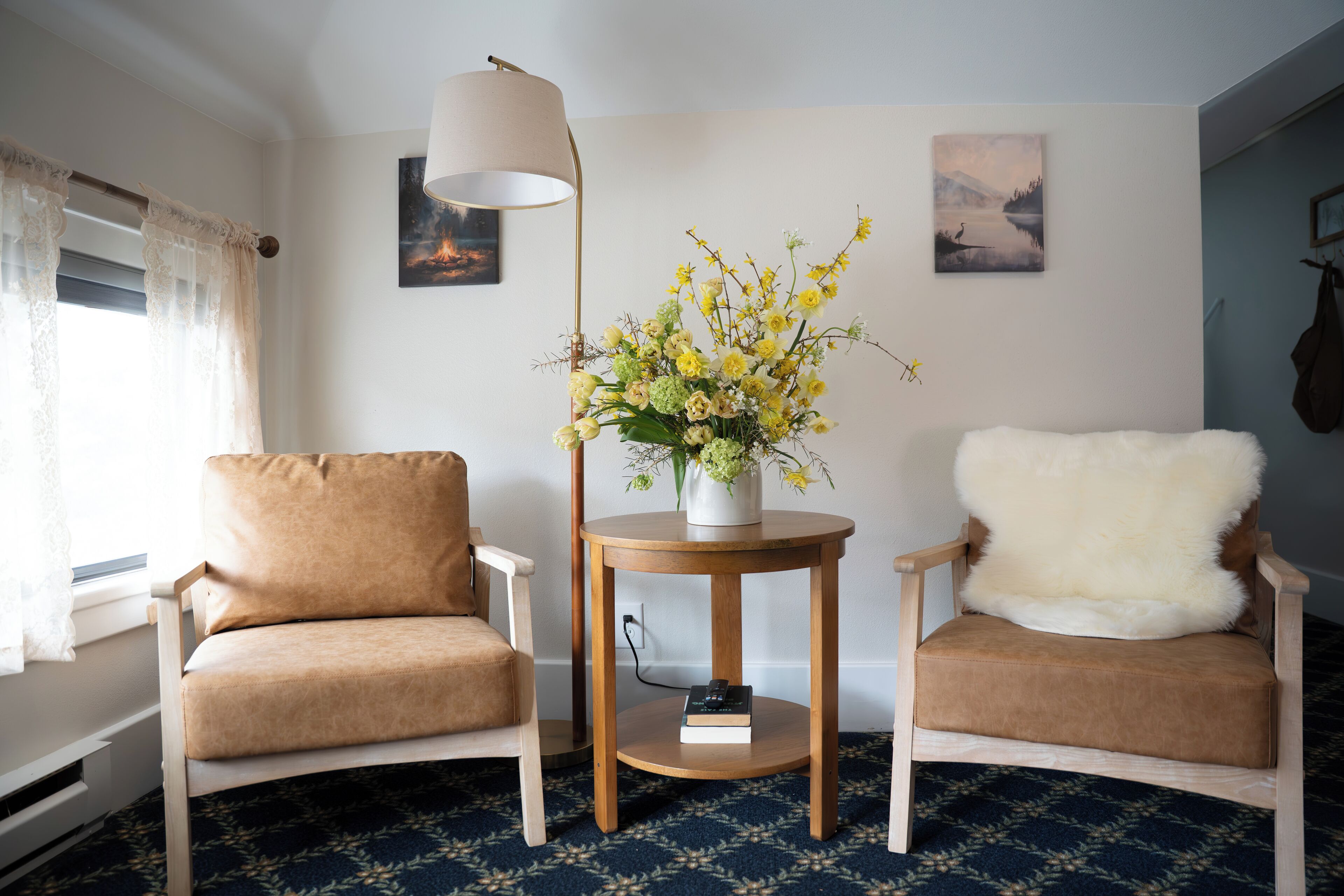 A room with two chairs, a table with a vase of flowers, a lamp, and artwork on the walls.