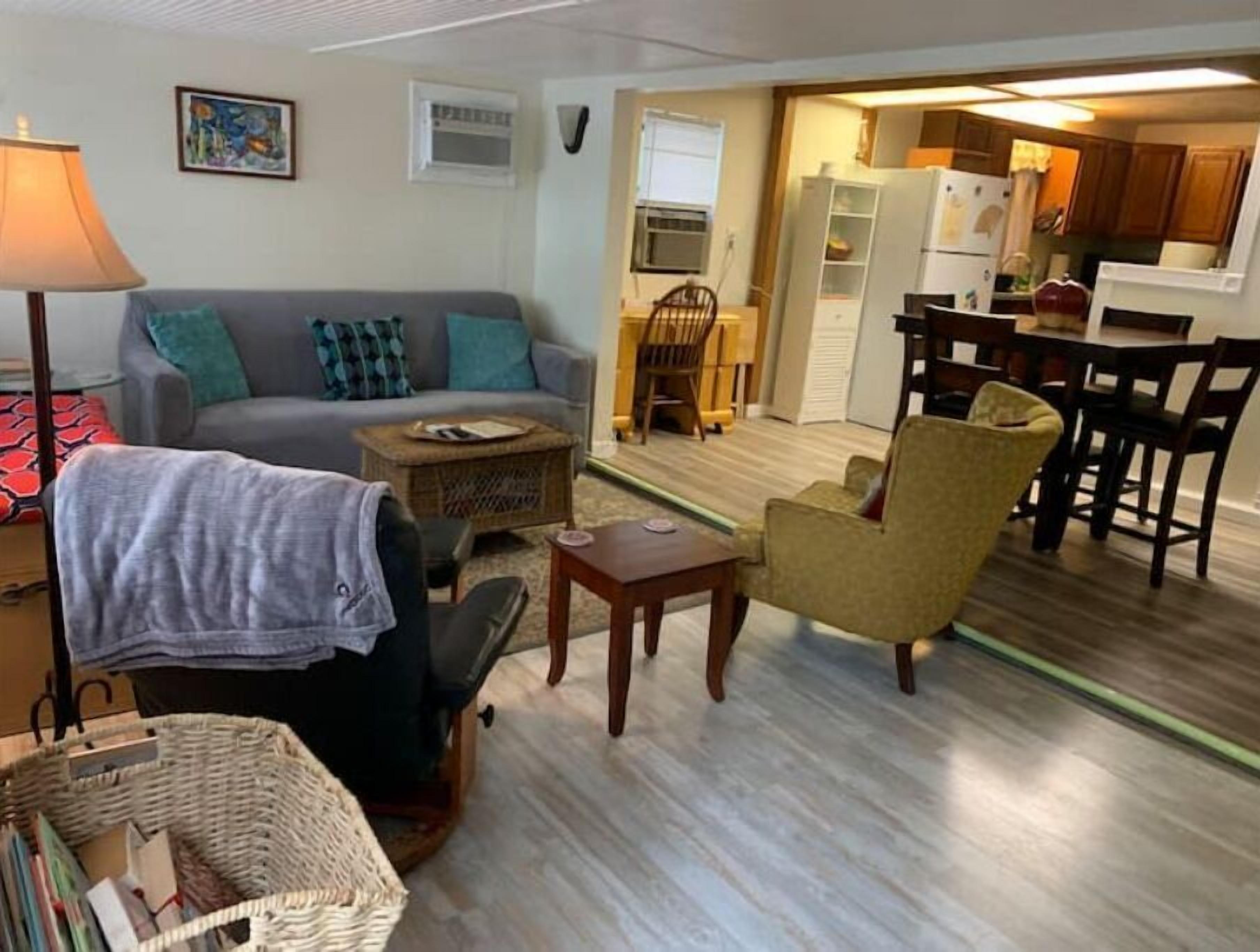 Una sala de estar con un sofá, un sillón y una mesita. Se observa un comedor con una mesa y sillas. Se ve una cocina al fondo.