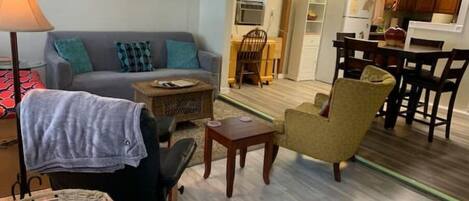 Una sala de estar con un sofá, un sillón y una mesita. Se observa un comedor con una mesa y sillas. Se ve una cocina al fondo.