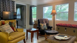 Una sala de estar acogedora con un sofá amarillo, un sillón negro, una mesita de madera y un otoman de mimbre.