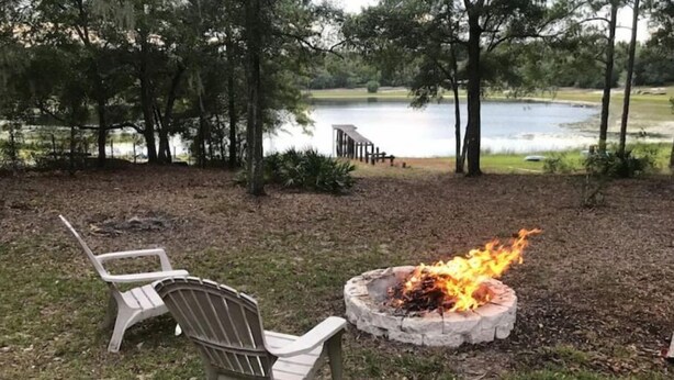 Un hogar con leña ardiendo, dos sillas de plástico blanco y una vista de un lago rodeado de árboles.