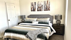 Una cama bien hecha con cubrecama a rayas y dos almohadas, flanqueada por dos mesitas de noche con lámparas y una obra de arte colgada en la pared.