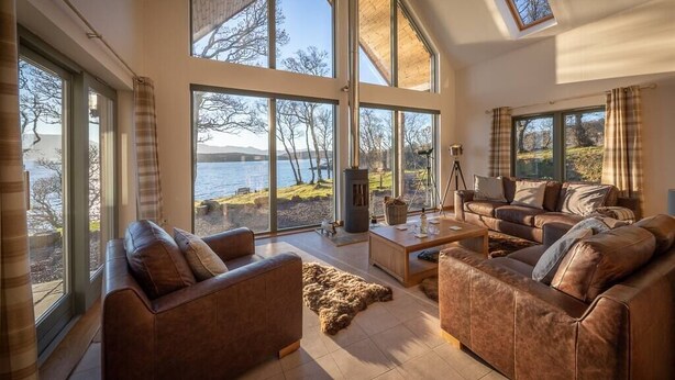 A spacious living room with large windows overlooking a lake, furnished with leather sofas and a coffee table. - The View, Duisdale Mor,  Sleat (Isle of Skye)