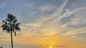 A balcony with two white lounge chairs, a palm tree, and a view of a cityscape at sunset. - 4 Bedroom House in Sotogrande Alto (San Roque)