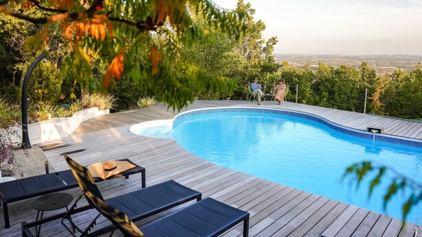 Una piscina con cubierta de madera, sillas de descanso y vista a la ciudad al fondo.