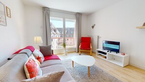Un salon moderne avec un canapé, un fauteuil rouge, un téléviseur et une fenêtre donnant sur des immeubles.