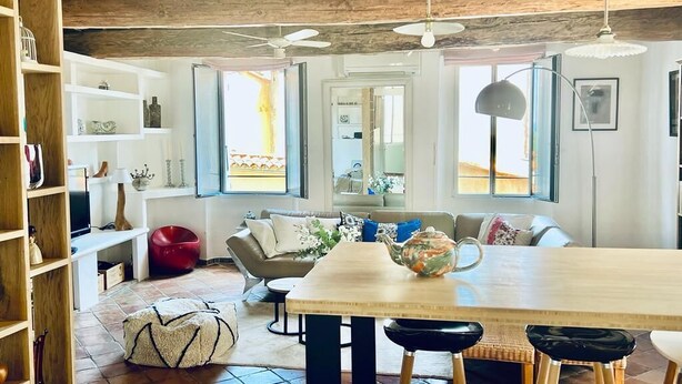 Een ruime woonruimte met een houten plafond, een grote houten tafel en een comfortabele bank. Er zijn meerdere ramen die zorgen voor natuurlijk licht en uitzicht op het omliggende landschap.