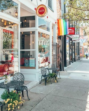 Un escaparate con un letrero que dice 'TASTE WINE', terraza con sillas y mesas de metal, y acera con diversos comercios.