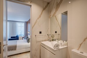 Quarto de hotel moderno com cama, banheiro com bancada de mármore e um espelho grande.