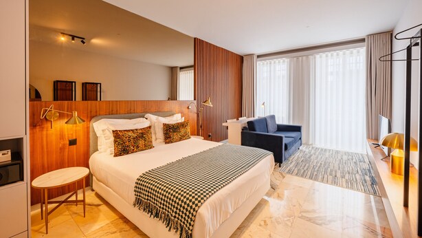 A modern hotel room with a large bed, a desk, a chair, and a view of the outdoors.