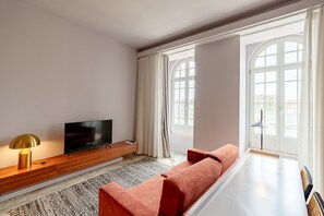 A modern living room with a flat-screen TV, wooden console, red sofa, patterned rug, and large windows with curtains.