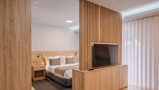 Una habitación de hotel moderna con una cama, un televisor montado en un panel de madera, una mesita de noche y un gran ventanal con cortinas.
