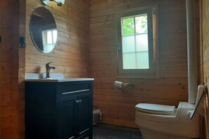Een houten badkamer in een blokhut, met een wastafel, spiegel, toilet en een raam.